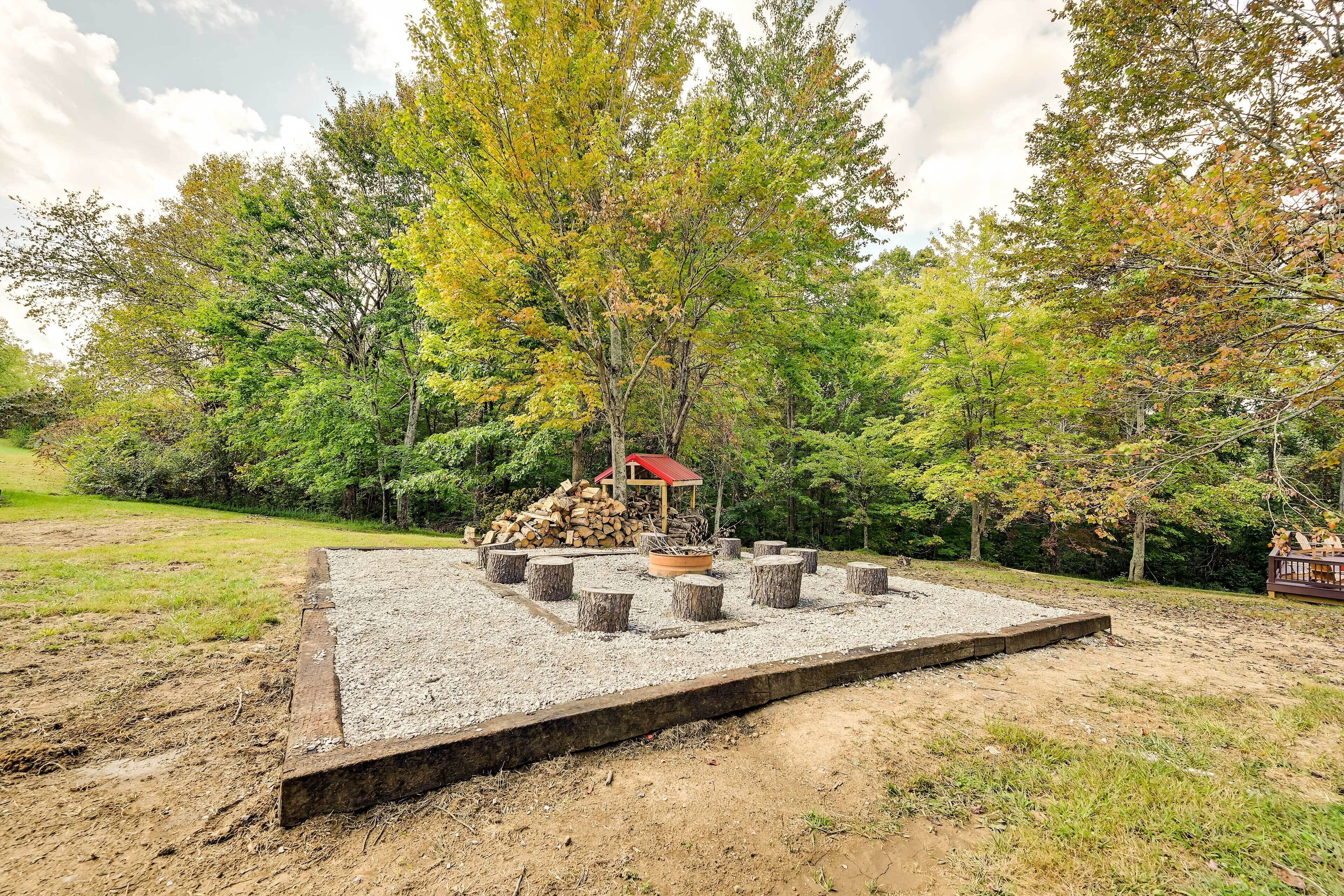 Une terrasse en gravier avec un foyer entouré de souches d’arbres, une pile de bois de chauffage et une structure surmontée d’un toit rouge.