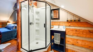 A bedroom with a glass-enclosed shower, wooden walls, a bed with blue bedding, and a small bathroom with a sink and towel rack.