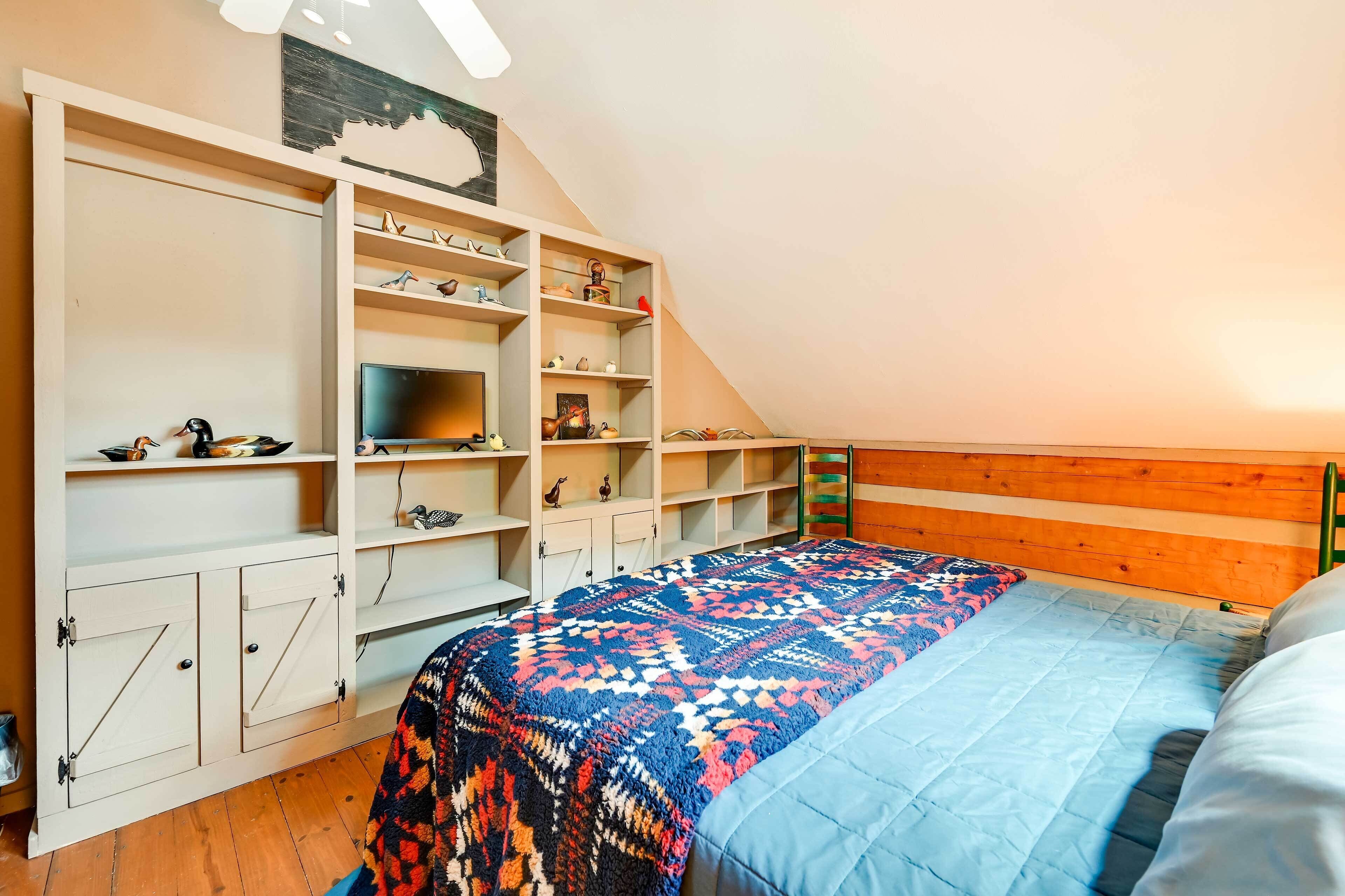 Une chambre avec un plafond en pente, un lit recouvert d’une couverture à motifs, une tête de lit en bois, des étagères intégrées où sont rangés divers objets, et un téléviseur.