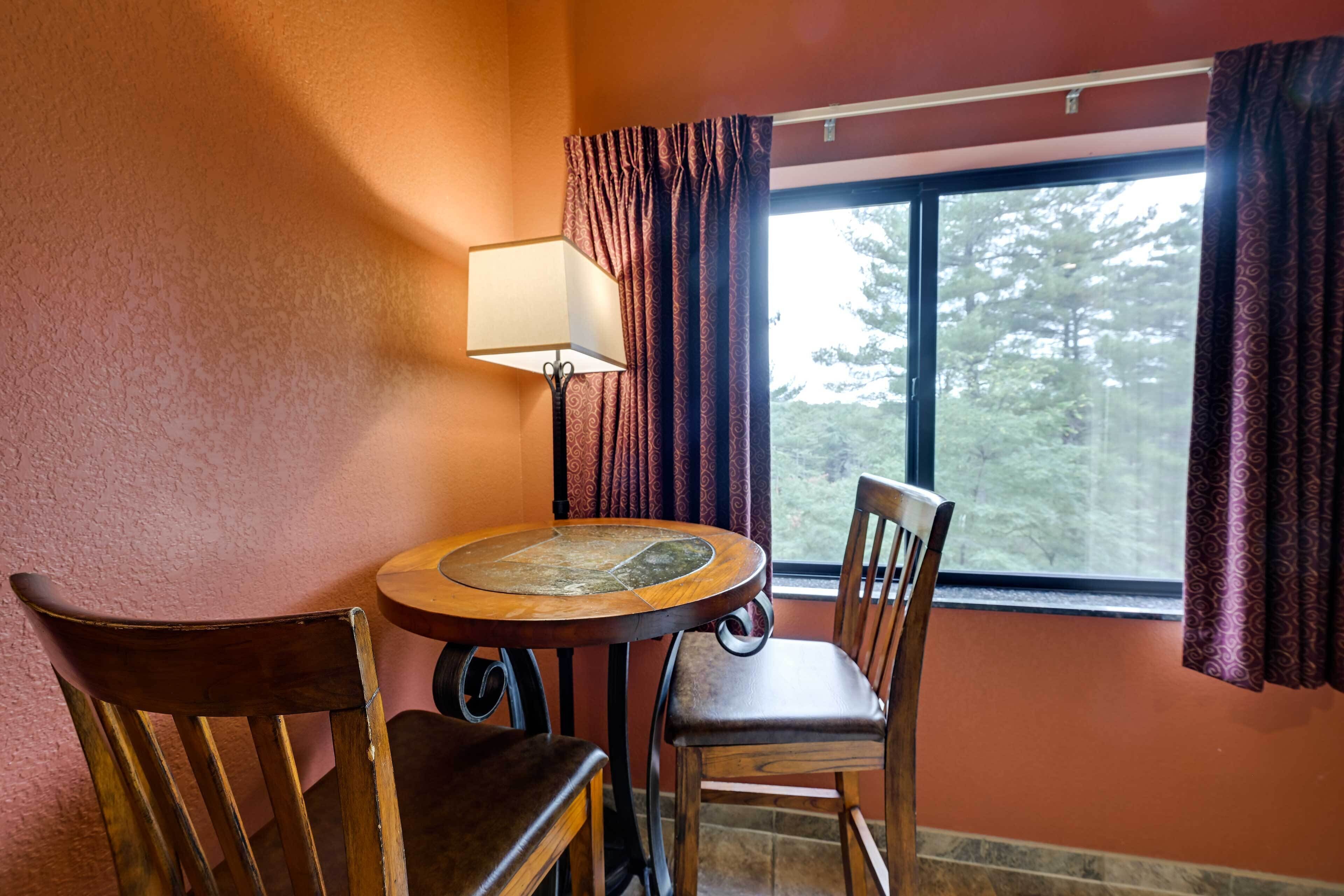 Une pièce avec une table ronde en bois, deux chaises en bois, une lampe et une fenêtre avec des rideaux.
