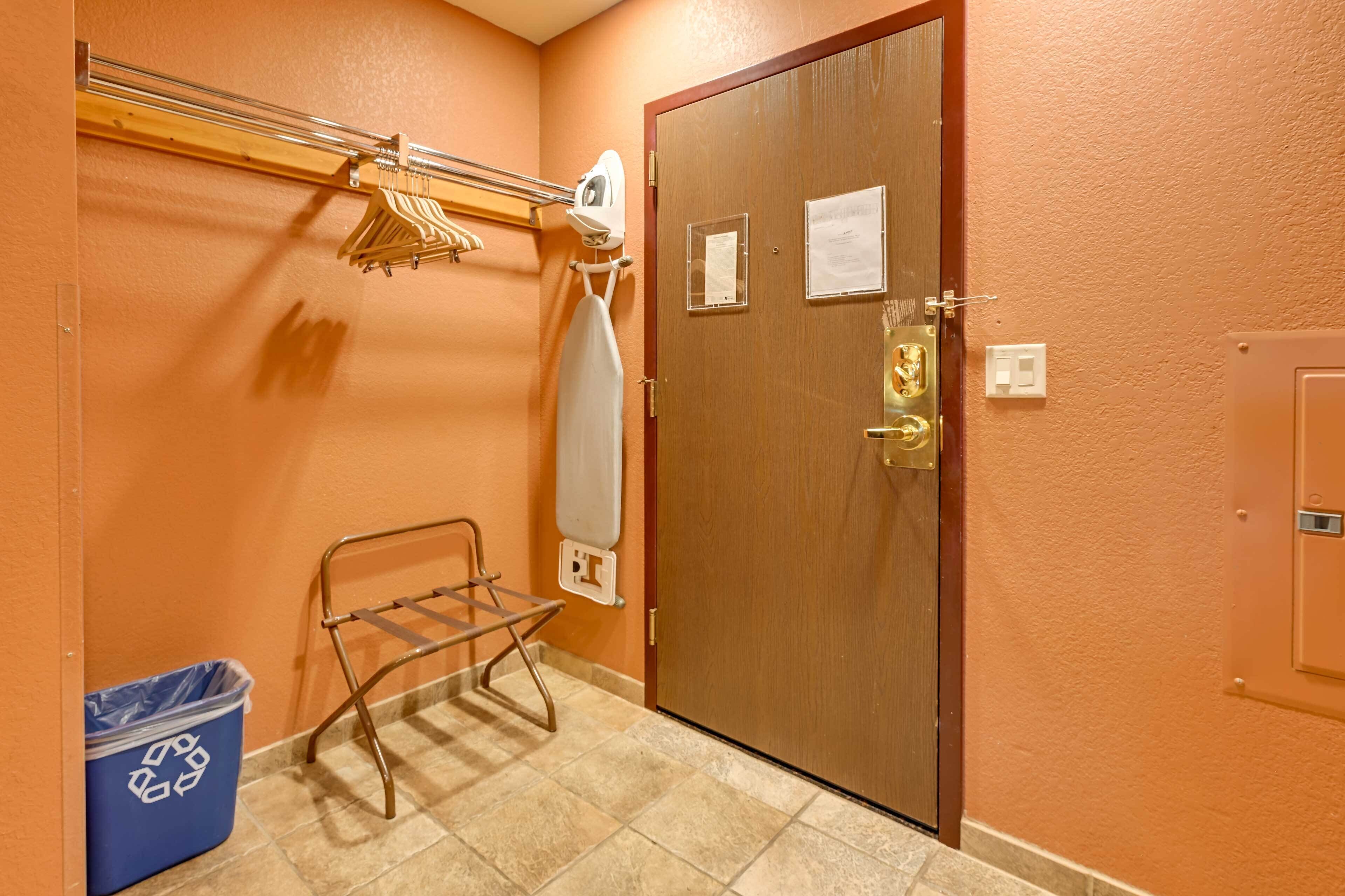 Camera d'albergo con una porta in legno, un asse da stiro fissata al muro, un cestino blu per la raccolta differenziata e un portasciugamani.