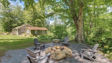 Una casa con terraza, rodeada de árboles, con una chimenea exterior y sillas Adirondack.