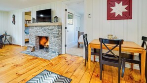 A cozy living room with a stone fireplace, wooden flooring, a dining table with chairs, and a mounted television. - 'mount Pine' Winter Retreat Near the Green Mtns! (Wardsboro)