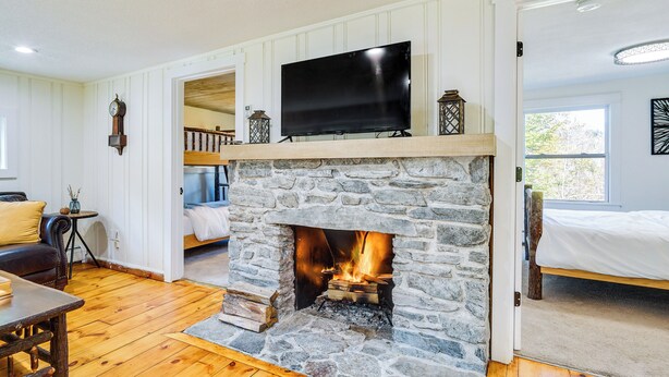 A stone fireplace with a flat-screen TV above it, a wooden floor, a bed with white bedding, and a view of the outdoors through a window. - 'mount Pine' Winter Retreat Near the Green Mtns! (Wardsboro)