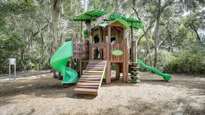 A wooden play structure with a slide and climbing elements, surrounded by trees and mulch.