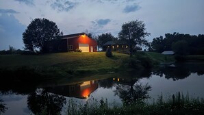 A modern house with a large garage, situated next to a calm pond, surrounded by trees and grassy areas.