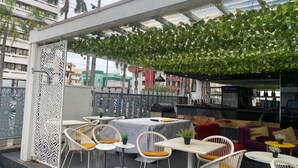 Un espace extérieur aménagé avec des chaises et des tables blanches, un coin bar agrémenté d’une enseigne Pepsi et une clôture décorative en métal.