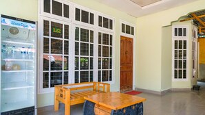 A room with a wooden bench and table, a Show Case refrigerator, and large windows with a number 122 sign. - OYO Life 2745 Nabeza House (Mandau)