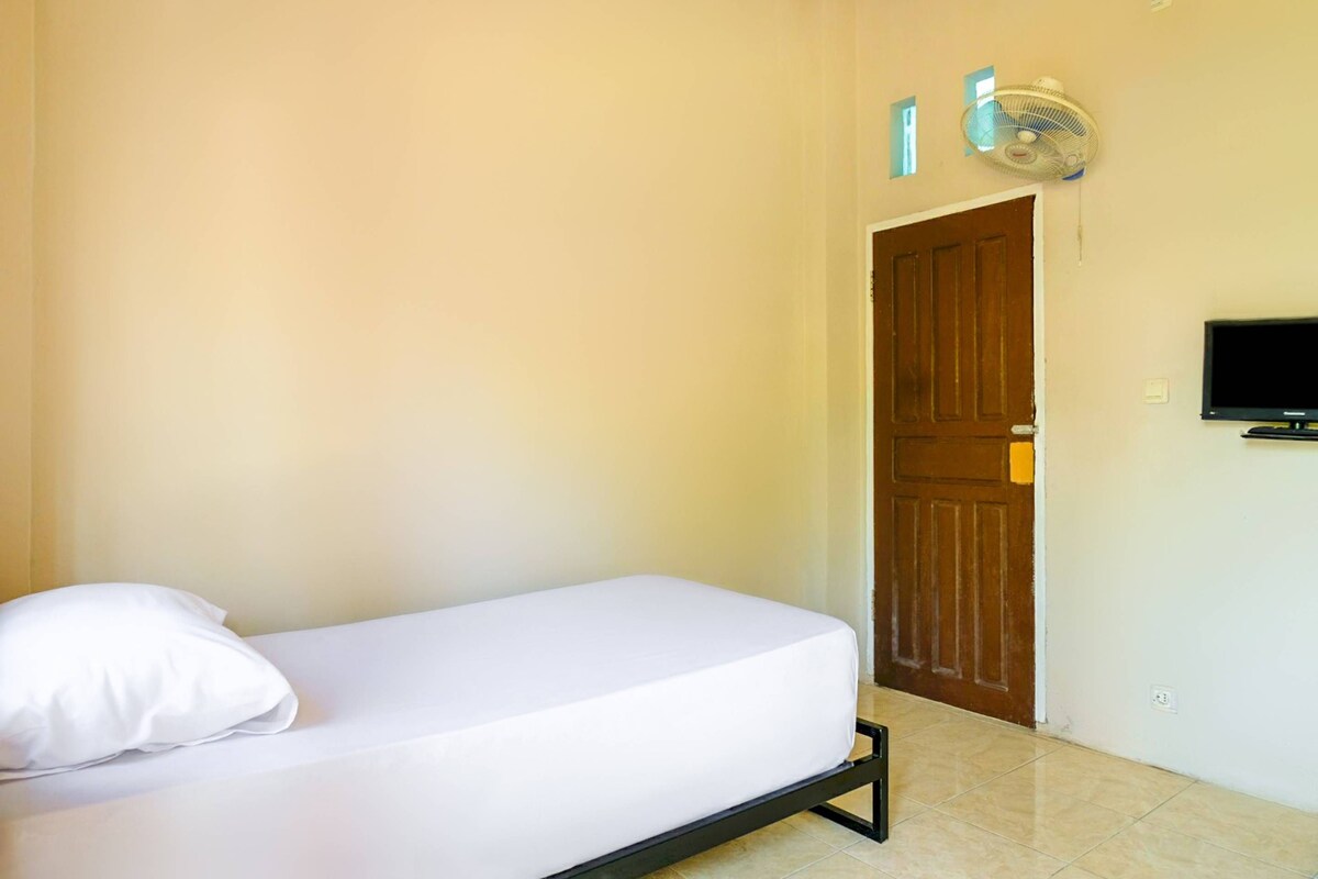 Una cama individual con colchón y almohada blancos, una puerta de madera, un televisor instalado en la pared y un ventilador de techo.