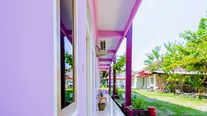 A corridor with a purple exterior, leading to a covered outdoor area with a garden and a red trash bin. - OYO Life 2745 Nabeza House (Mandau)