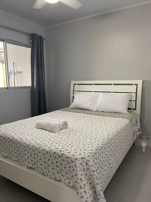 Un lit bien fait, recouvert d’une couvre-lit à motifs floraux et agrément é d’oreillers blancs, une fenêtre avec des rideaux et un ventilateur de plafond.