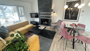 A modern living room with a sofa, coffee table, fireplace, TV, and dining area with chairs and a glass table. - Location! New, Modern Townhouse Steps from 124 Street! (Edmonton)