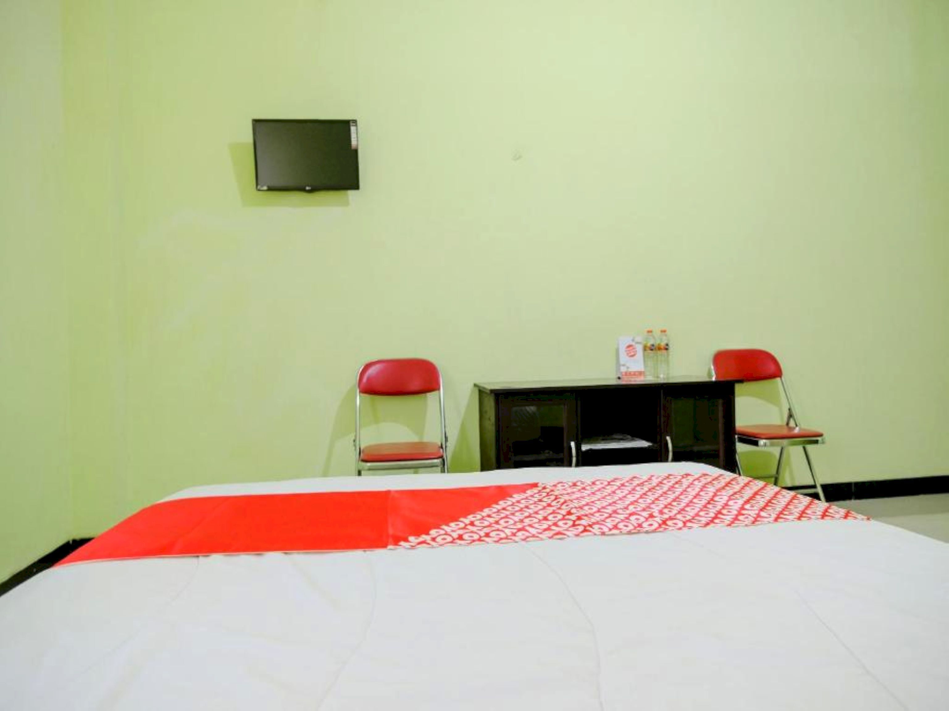 Una habitación con una cama, dos sillas rojas, una mesita con botellas y un televisor en la pared.
