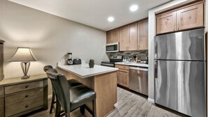 A compact kitchen with wooden cabinets, a stainless steel refrigerator, a microwave, and a bar stool.