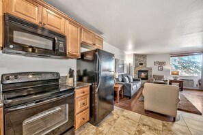 A modern kitchen with stainless steel appliances, wooden cabinets, and a stone fireplace in a living area with a sofa and a view of trees.