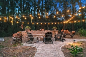 Un patio al aire libre con sillas tipo Adirondack, una chimenea de piedra y luces colgantes.