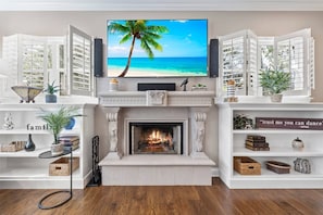 Una sala de estar con chimenea, un televisor mostrando una escena de playa y contraventanas abiertas.