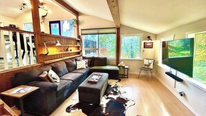 A cozy living room with a wooden ceiling, a large window, a flat-screen TV, and a leather sofa.