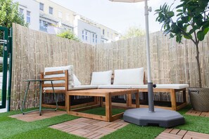 A wooden outdoor seating area with a bench and a table, surrounded by a bamboo fence and artificial grass.