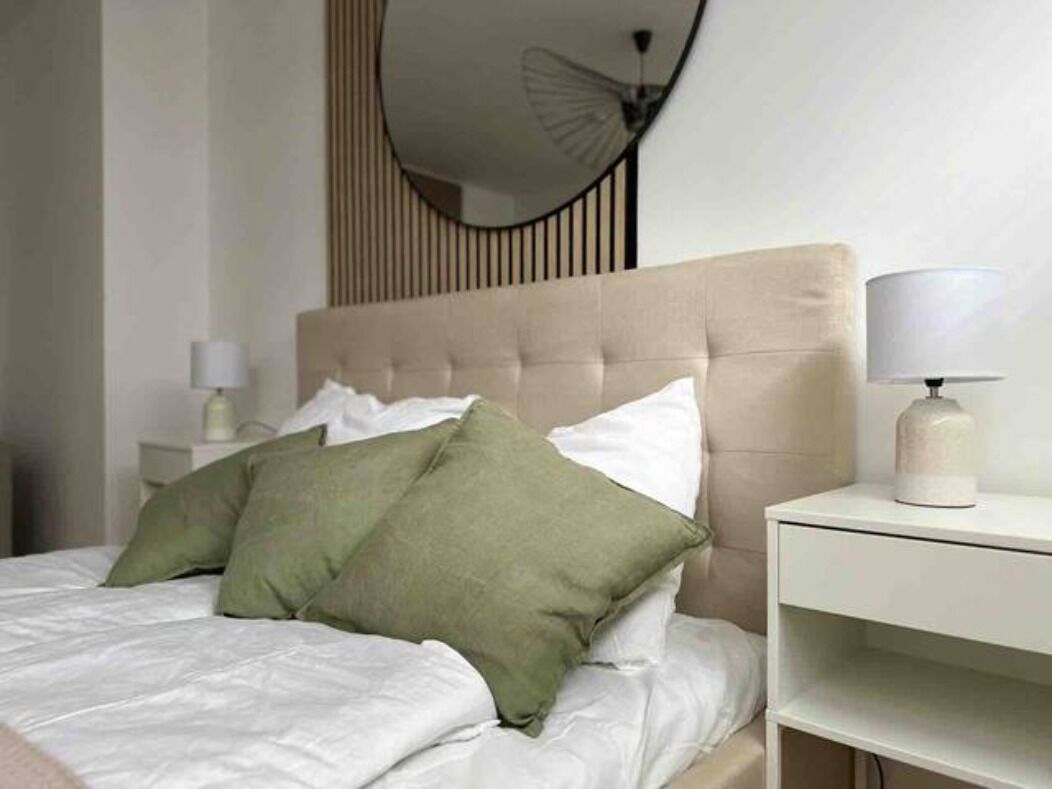 Een netjes opgemaakt bed met witte en groene kussens, een beige hoofdbord en een nachtkastje met een lamp.