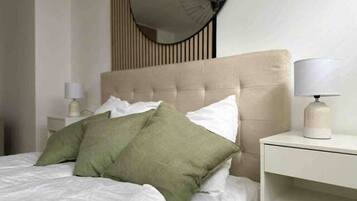 Een netjes opgemaakt bed met witte en groene kussens, een beige hoofdbord en een nachtkastje met een lamp.