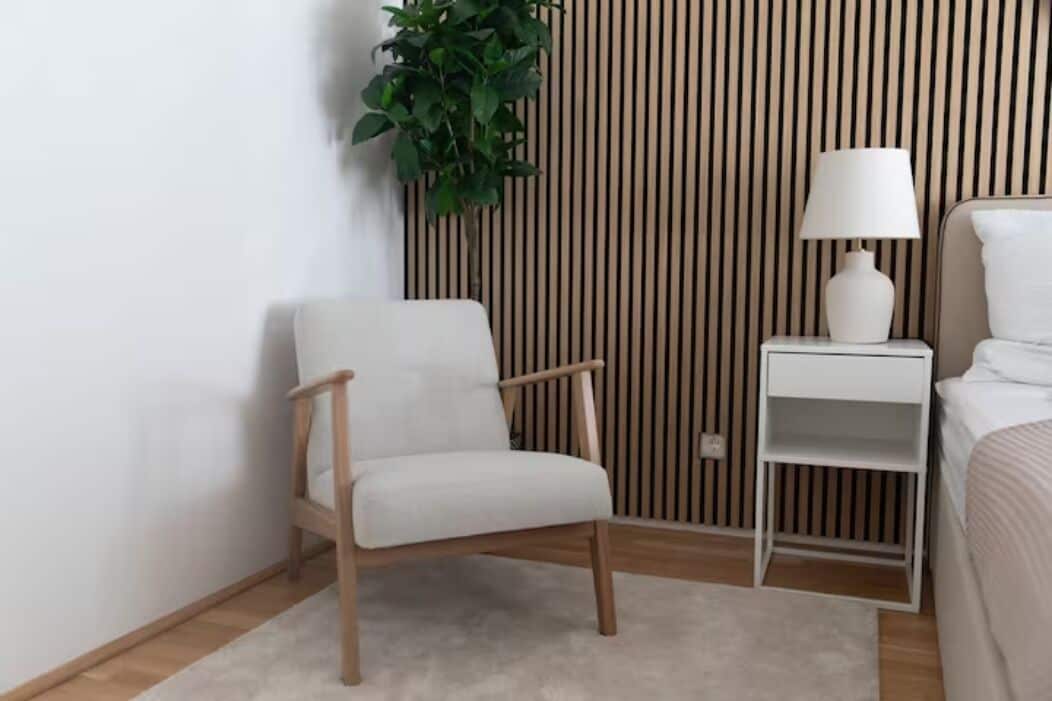 Een moderne slaapkamer met een wand van houten latten, een witte stoel, een nachtkastje met een lamp, en een bed met wit beddengoed.