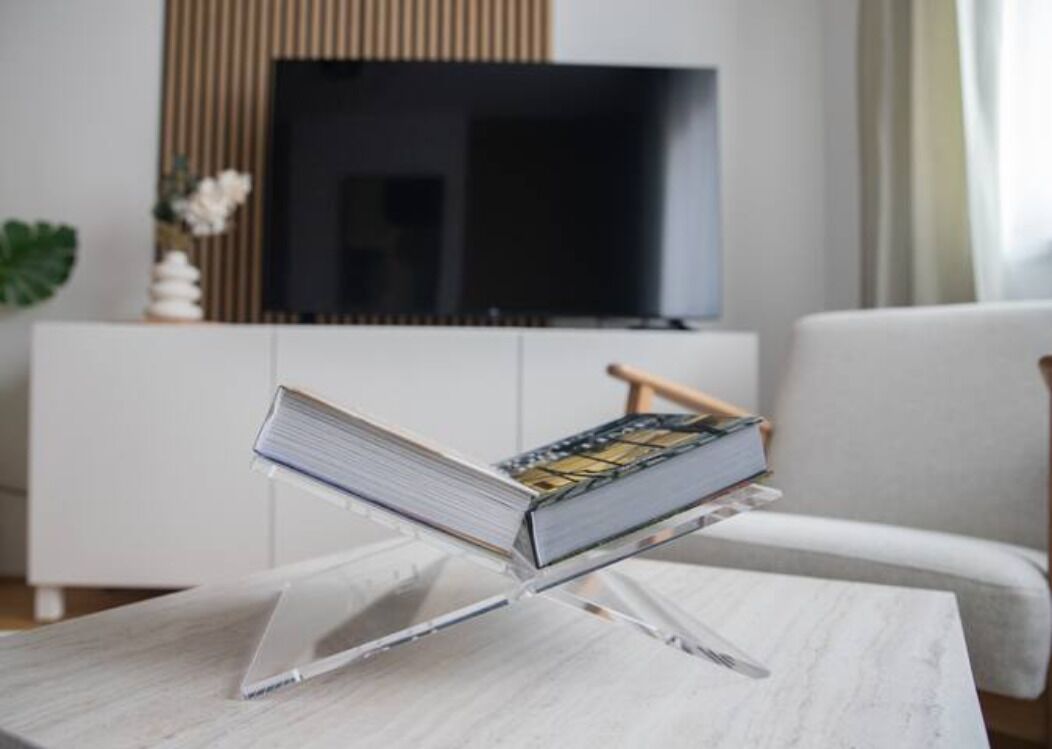 A book on a clear stand on a marble table in a modern living room with a TV and a white sofa.