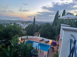 Zona de piscina con sillones de descanso y vista a un paisaje urbano al atardecer.