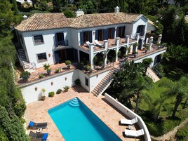 Una casa grande y blanca de dos pisos, con techo de tejas, piscina y varios balcones.