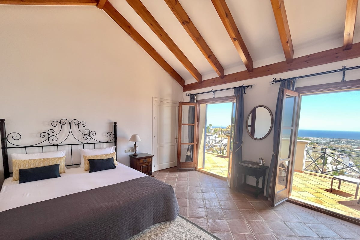 A bedroom with a large bed, a wooden ceiling, and a balcony with a city view.