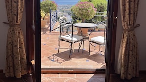 An open sliding glass door leading to an outdoor patio with a table and chairs, a view of the sea, and a balcony with a railing.