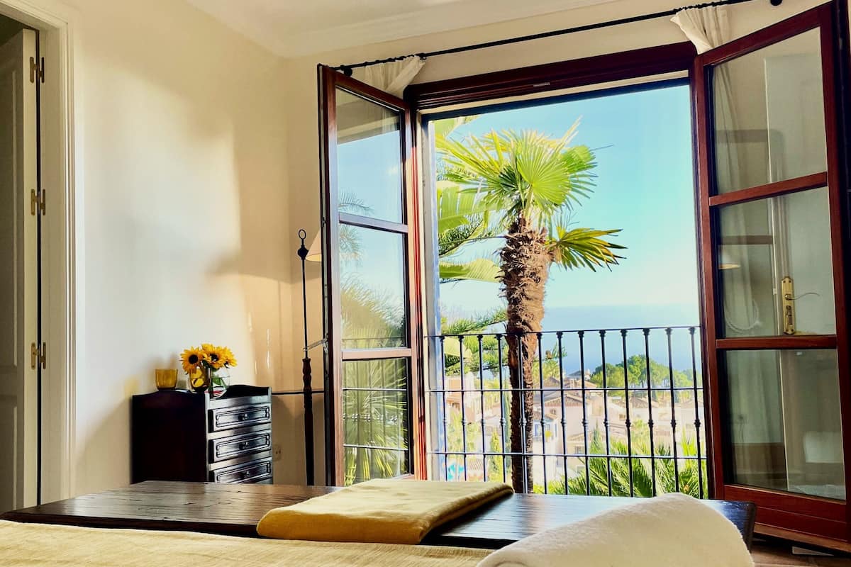 A bedroom with a large window, a bed with a rolled towel, a nightstand with a lamp, and a view of palm trees and buildings.