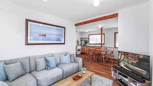 Una sala de estar moderna con un sofá gris, una mesa de centro de madera y un televisor de pantalla plana. También se aprecia un comedor con sillas de madera y una cocina al fondo.