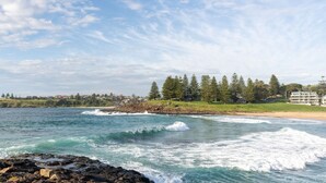 A coastal view with waves crashing onto a rocky shore, a sandy beach, and a row of buildings in the background. - Blue Horizon on Manning (Kiama)