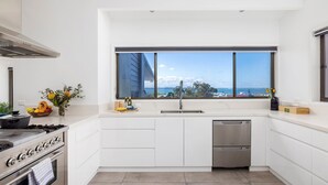 Una cucina moderna con mobili bianchi, piano cottura in acciaio inossidabile, lavastoviglie e un'ampia finestra con vista sul mare.