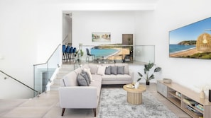 Una sala de estar moderna con un sofá, una mesa de centro y una estantería. Se ve una escalera que sube a un nivel superior con barandilla de vidrio.