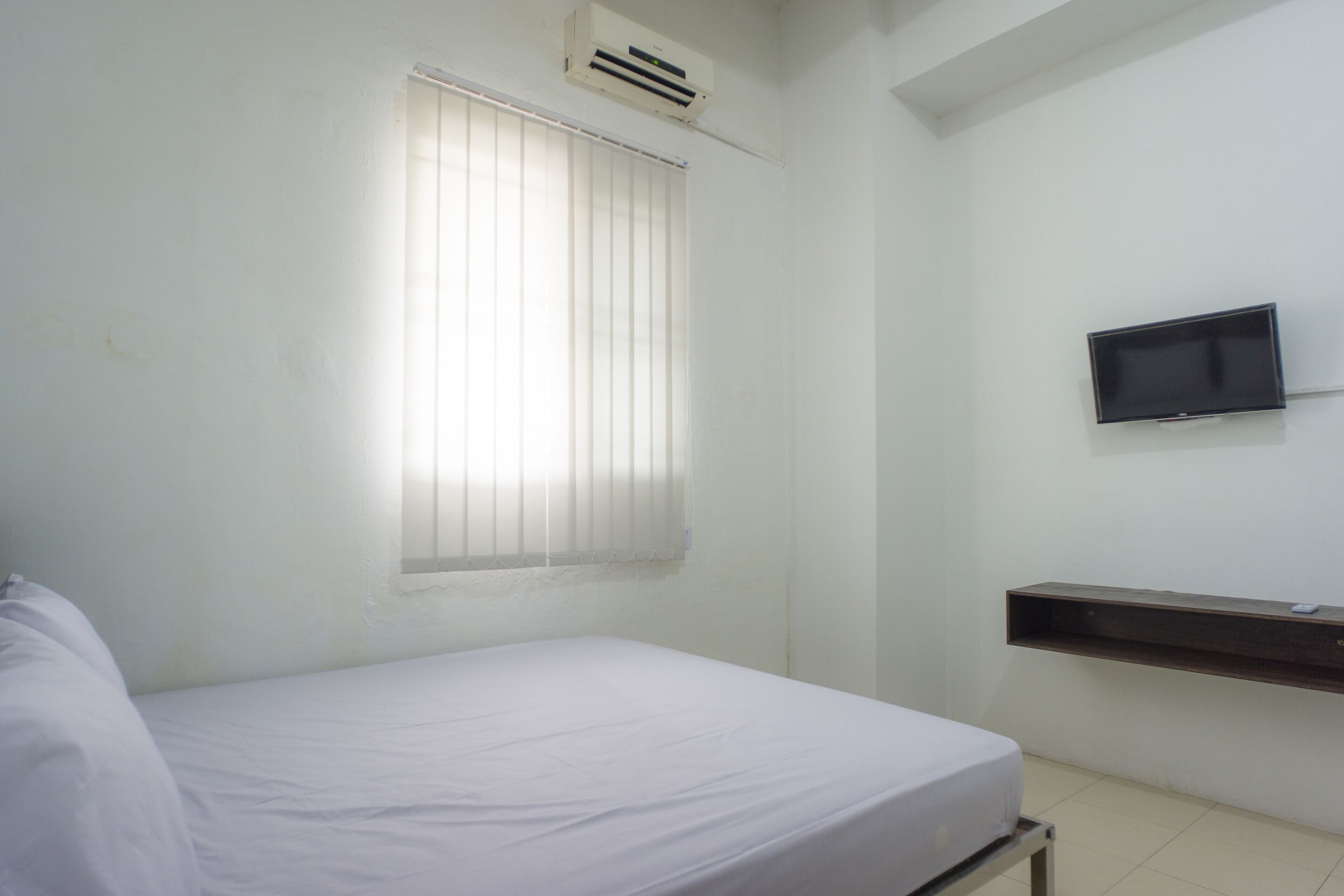 A small, clean room with a single bed, a mounted TV, and a window with blinds.