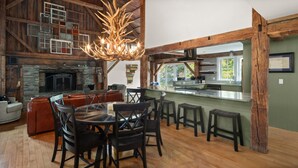 A rustic interior with a stone fireplace, wooden beams, a dining area, and a kitchen with a large window.