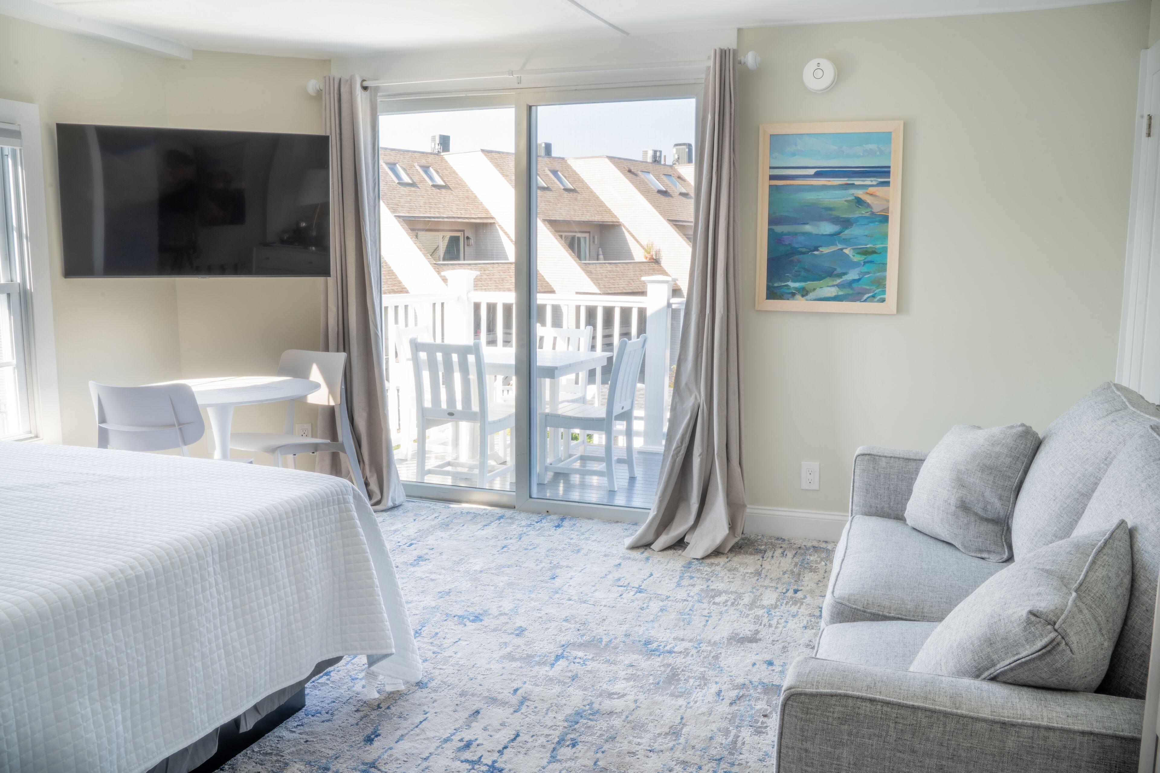 A hotel room with a bed, a grey sofa, a TV mounted on the wall, a painting, and a balcony with a view of buildings.