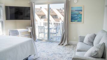 A hotel room with a bed, a grey sofa, a TV mounted on the wall, a painting, and a balcony with a view of buildings.