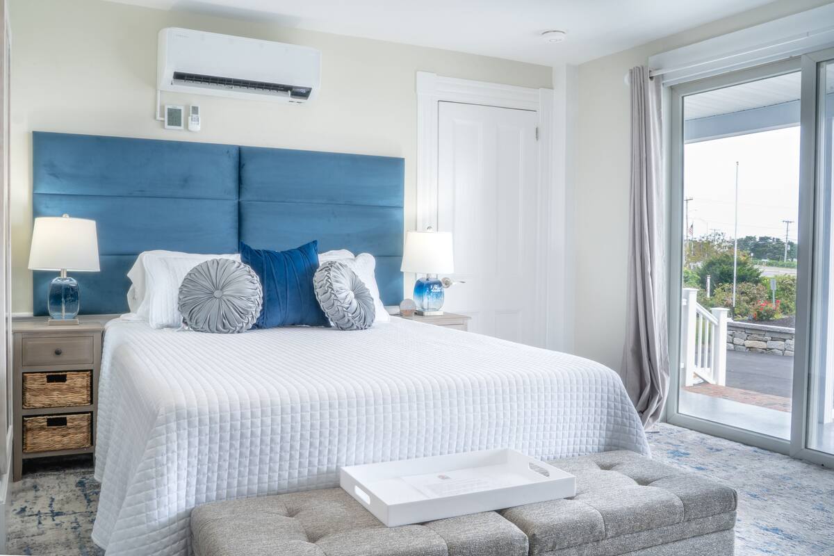 A hotel room with a large bed, two bedside tables with lamps, a view of the outdoors through a large window, and an air conditioning unit on the wall.