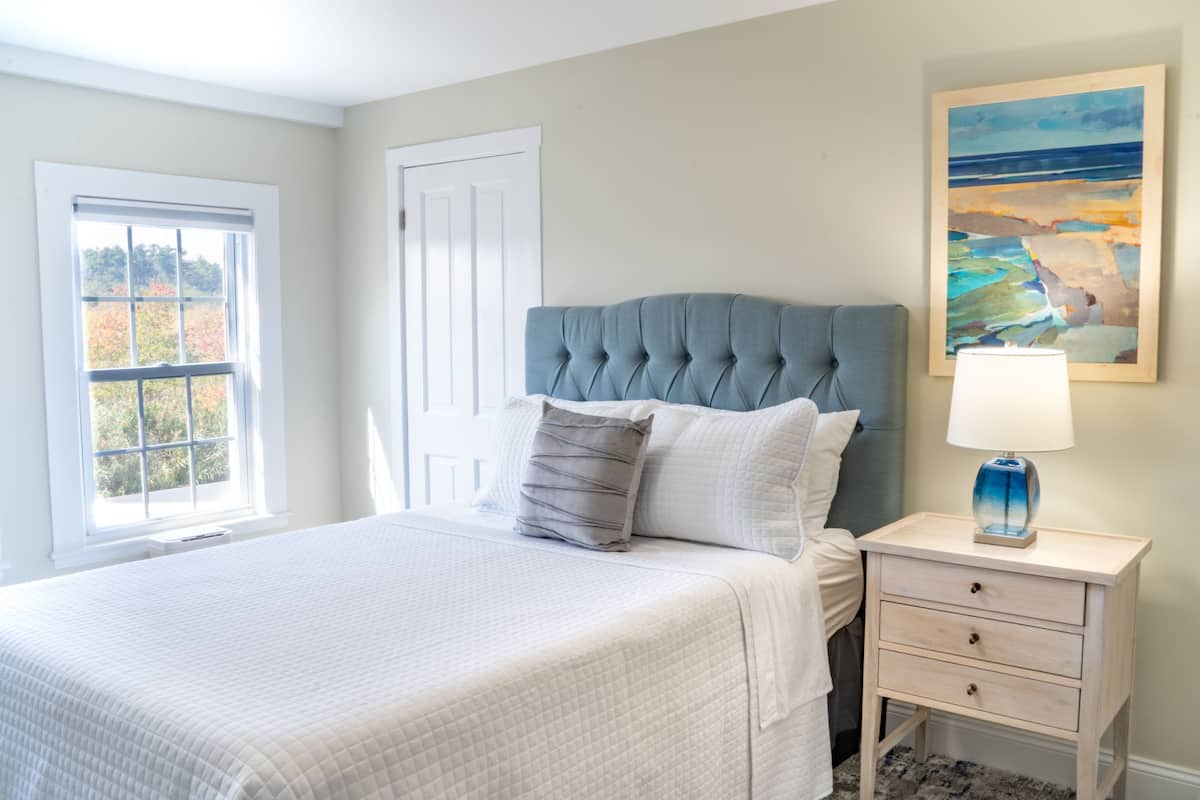 A bedroom with a large bed, a nightstand with a lamp, a window with a view of trees, and a painting on the wall.