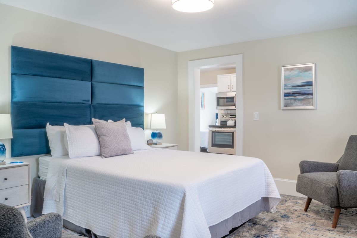 A bedroom with a large bed, a grey armchair, a nightstand, and a wall-mounted lamp.