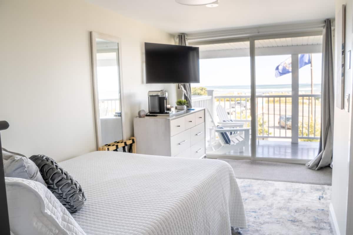 A bedroom with a bed, a dresser, a television, and a balcony with a view of the beach.