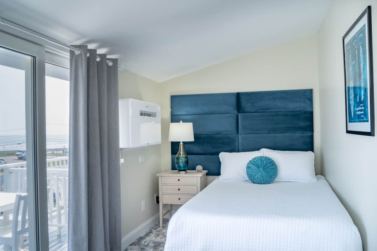 A hotel room with a large bed, a nightstand, a lamp, and a view of the ocean.