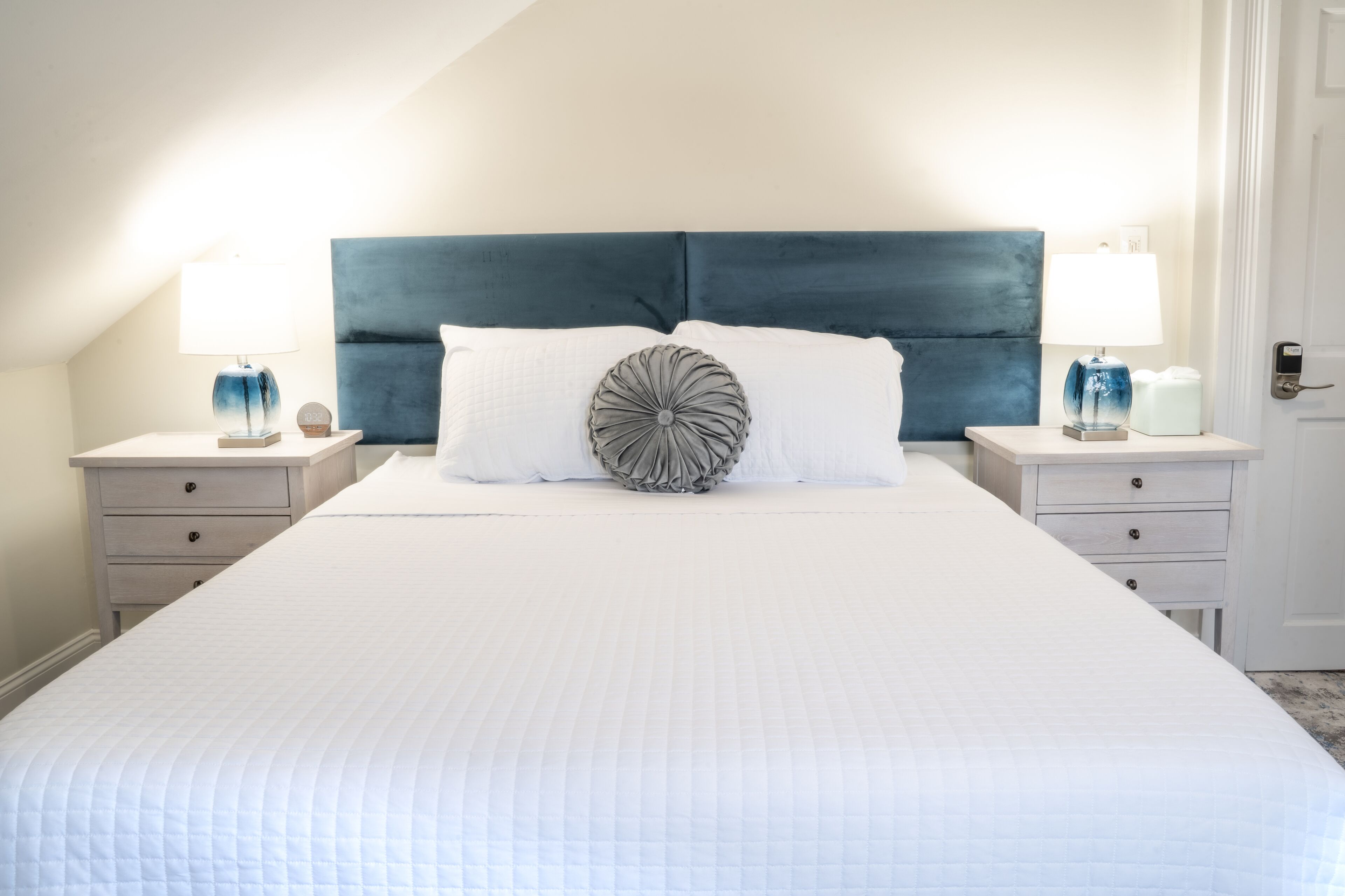 A neatly made bed with a quilted white duvet, a dark blue upholstered headboard, and two bedside tables with lamps and decorative items.