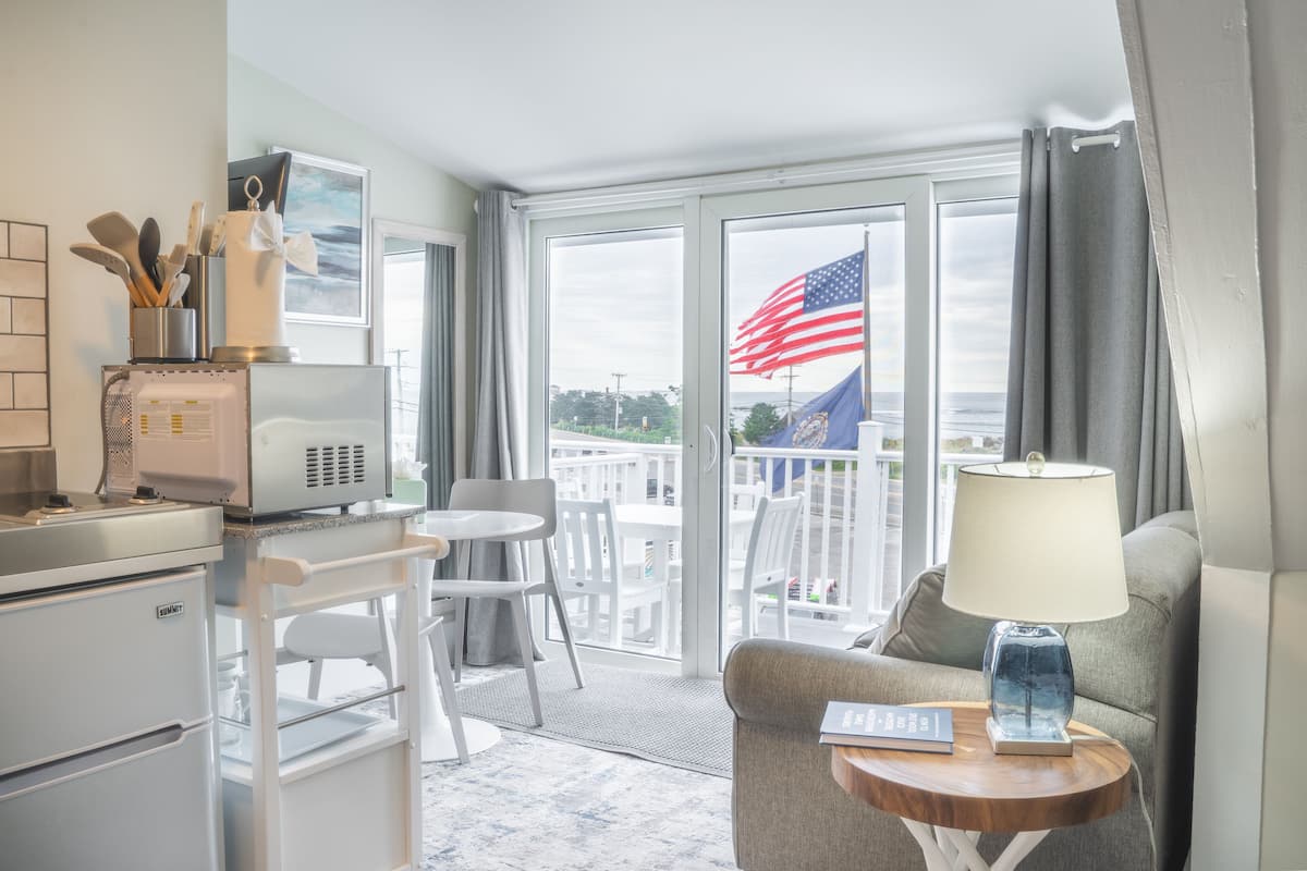 A hotel room with a kitchenette, a dining area with chairs, a sofa with a lamp, and a balcony with an American flag.