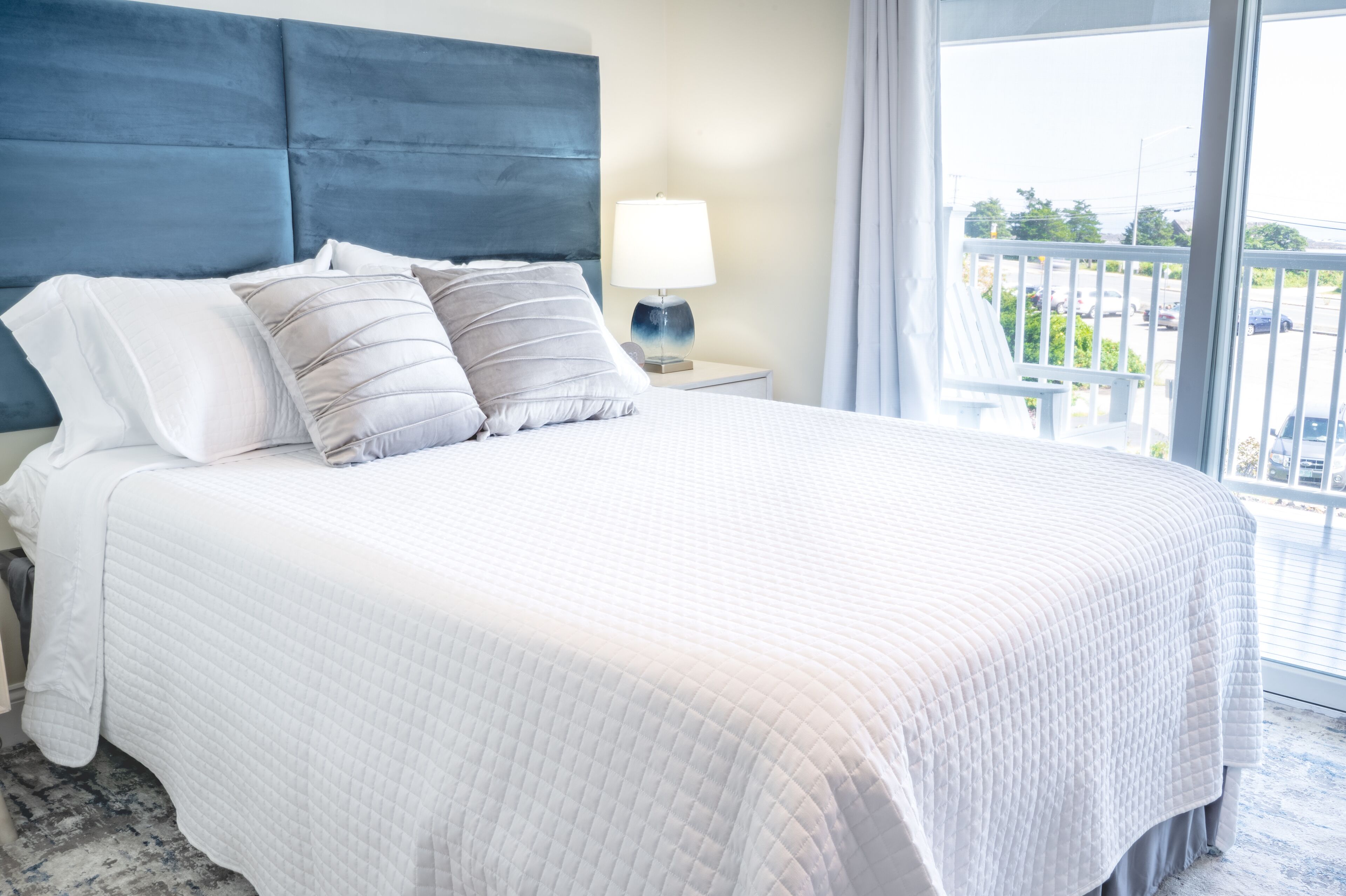 A neatly made bed with a white quilted bedspread and two pillows, a bedside table with a lamp, and a view of a balcony with a railing and greenery.