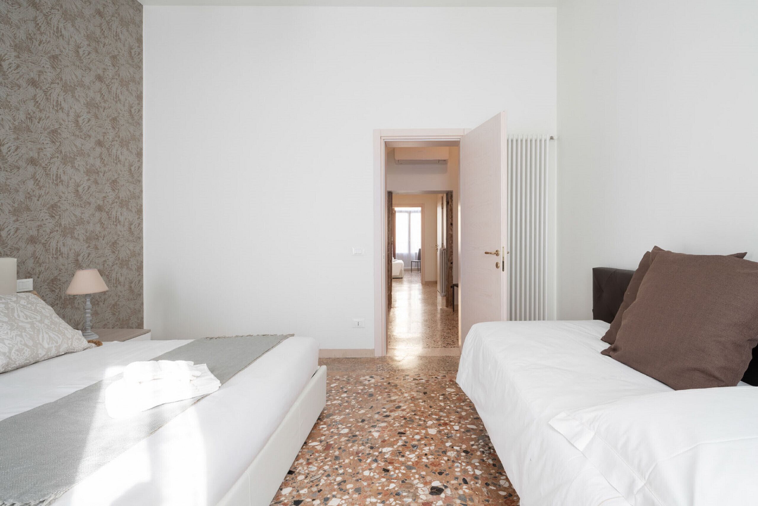 Hotelkamer met een bed, nachtkastje met lamp, radiator en deur naar een andere kamer.