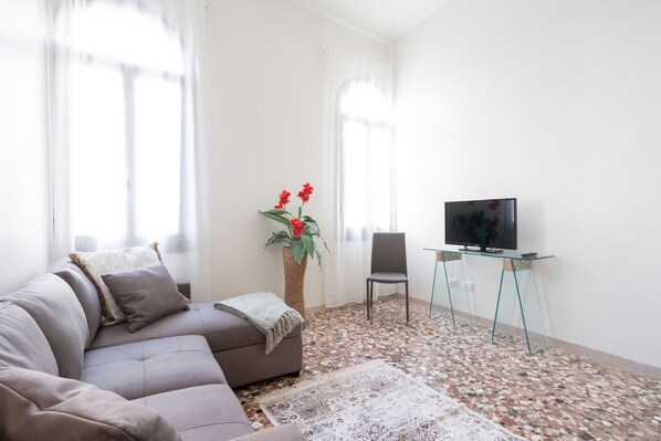 Een woonkamer met een bank, een glazen salontafel, een televisie, een stoel en een decoratieve plant.